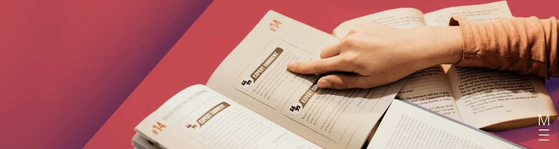 Hand pointing to text in an open book, surrounded by study materials
