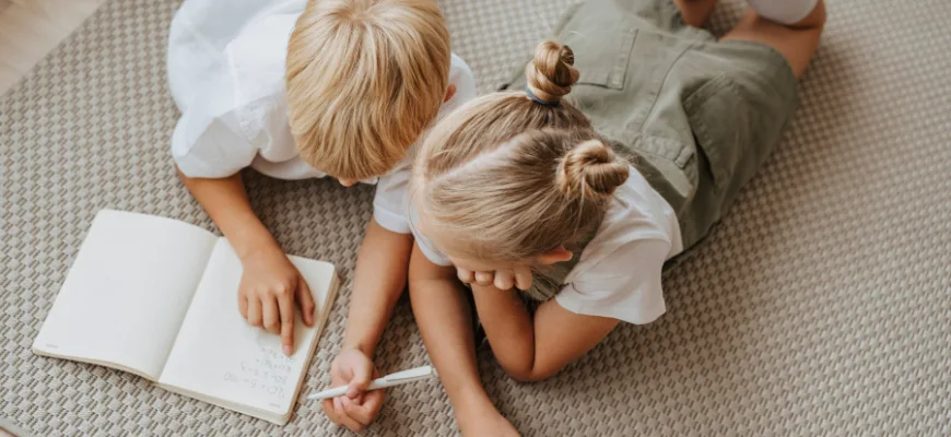 matrix education Two children lying on a carpet, collaborating on homework in an open notebook. One child points to the page while the other holds a pen.