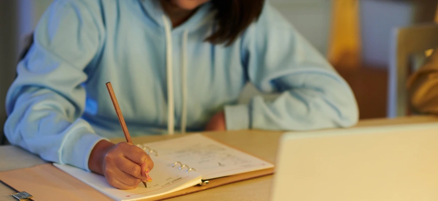 A student in a blue hoodie writing notes in a notebook while studying at a desk