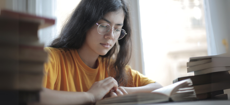 Student reading a book at a desk surrounded by stacks of books.