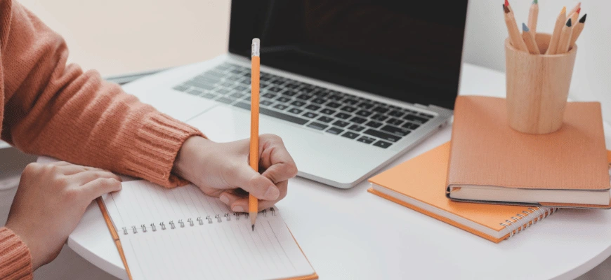Person writing in a notebook with a laptop on the desk