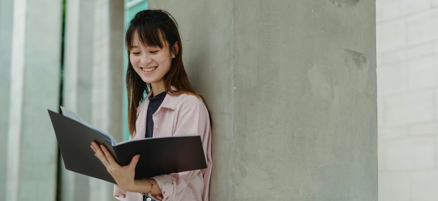 A smiling student leaning against a concrete wall, reading a black folder.