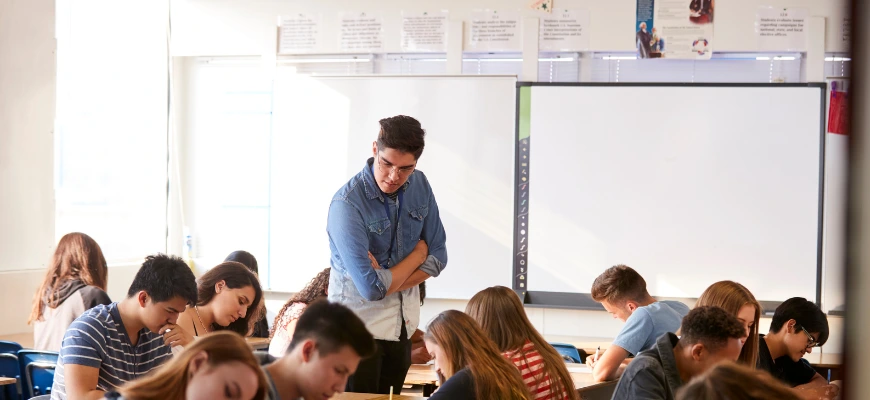 A classroom of students focused on their work, with a teacher standing and observing