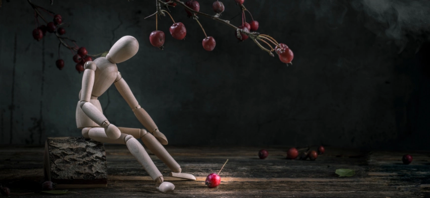 Wooden mannequin sitting on a block, gazing at a fallen cherry - representing gravity