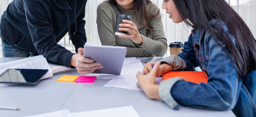 Three students collaborating at a table with notes, a tablet, and coffee cups.